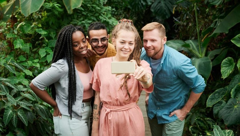 Four friends smiling and taking a selfie amidst lush greenery, capturing a joyful moment outdoors.